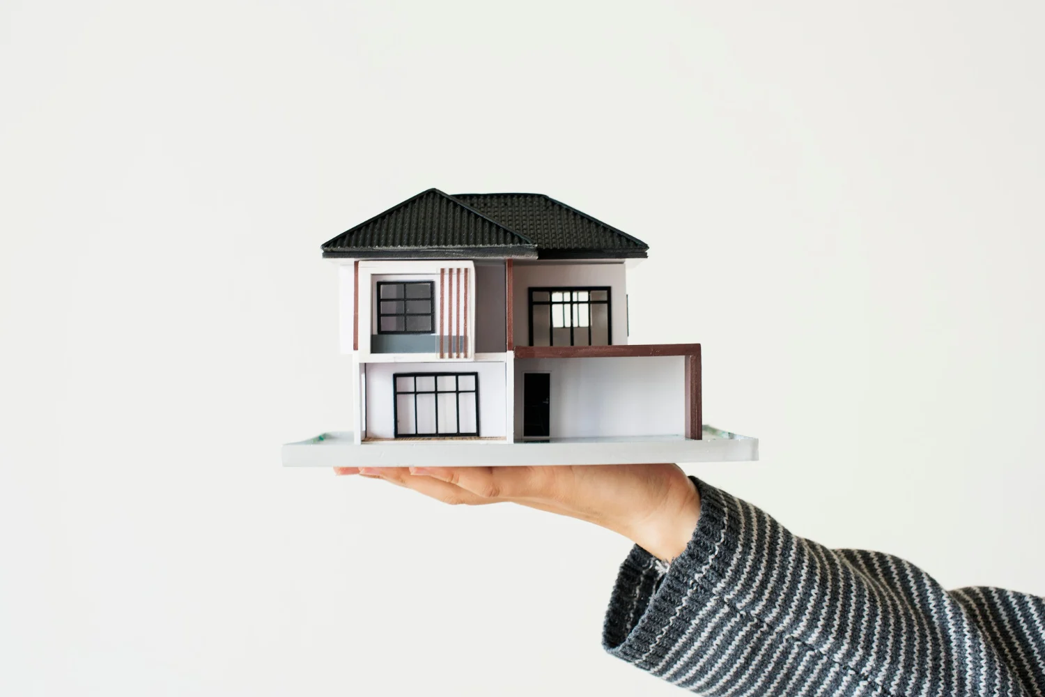 A person holds a miniature model of a modern two-story house on a flat base, with a clean white background. The model features a black roof, large windows, and a garage, symbolizing homeownership or real estate concepts.