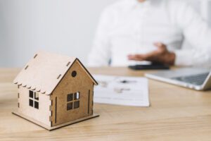 A miniature wooden house sits on a desk in front of a businessman, with paperwork and a laptop in the background. The image conveys a conversation or consultation about real estate or mortgage terms.