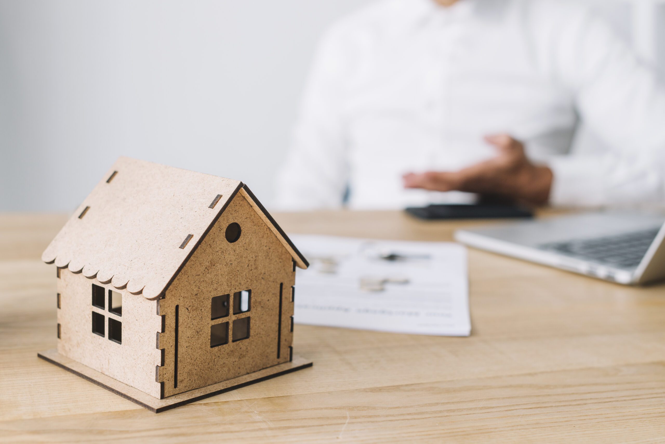 A miniature wooden house sits on a desk in front of a businessman, with paperwork and a laptop in the background. The image conveys a conversation or consultation about real estate or mortgage terms.