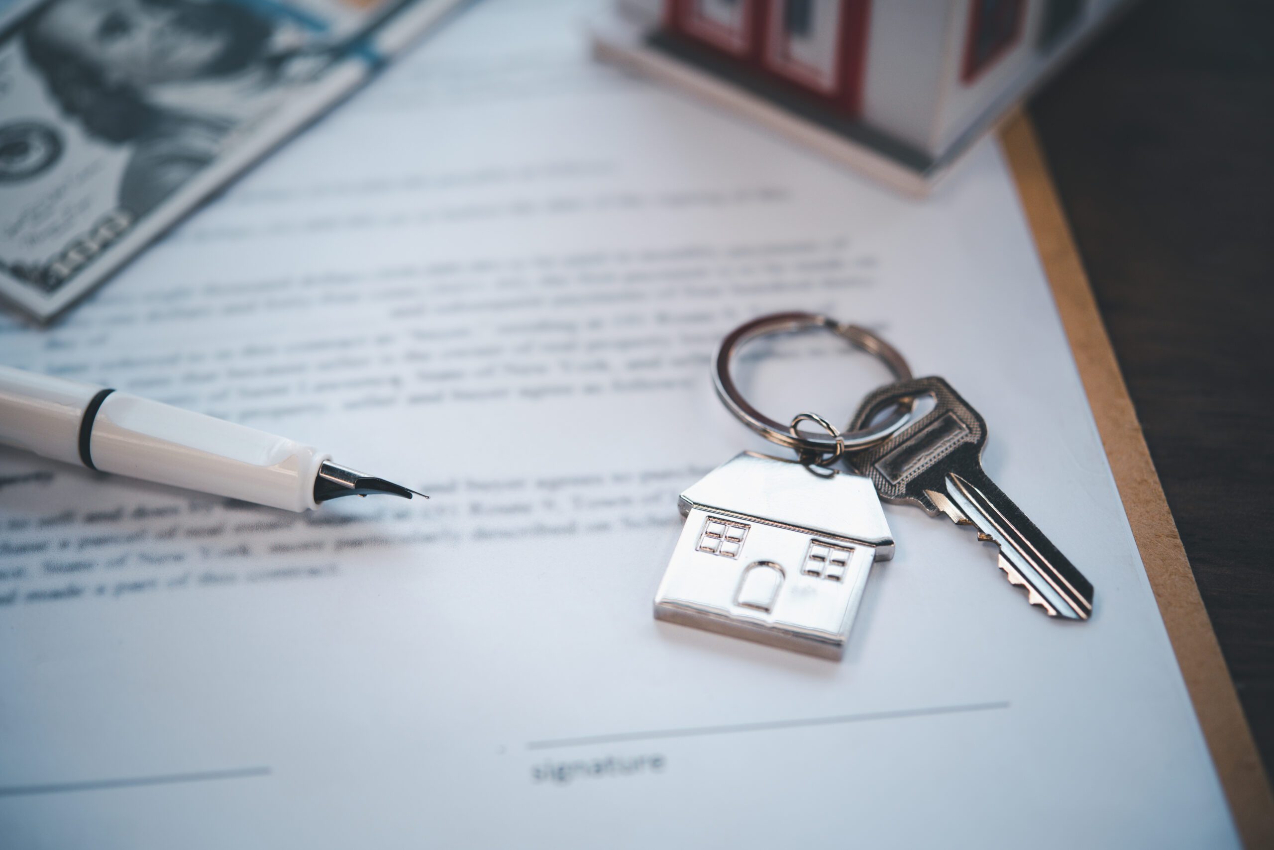 A close-up of a house key with a house-shaped keychain resting on top of a contract, next to a fountain pen. The setup suggests a home sale or rental agreement ready to be signed.