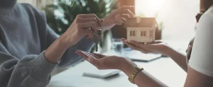Two women exchange a key and a miniature house, symbolizing a home purchase or real estate transaction. The scene takes place in a well-lit, professional setting, suggesting a successful property handover.